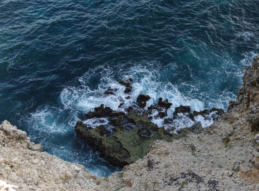 A wave crashes against a rocky shore, with crystal blue water visible in the background. This image is part of the post about how ocean currents affect climate.